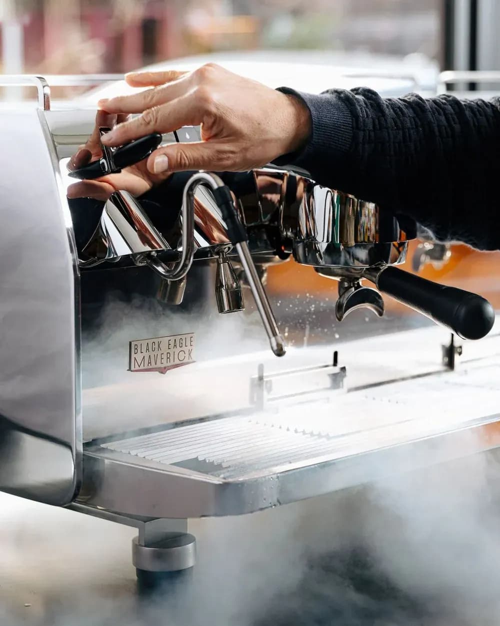 A hand adjusting the steam wand on a white Black Eagle Maverick, the badge visible beside a portafilter, steam rising.