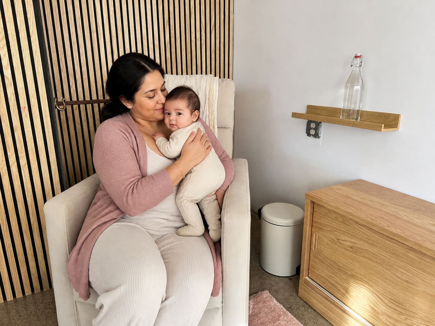 A mama cuddling her baby close in a cream recliner in the Mama Space, with a fresh bottle of water on the shelf beside her.