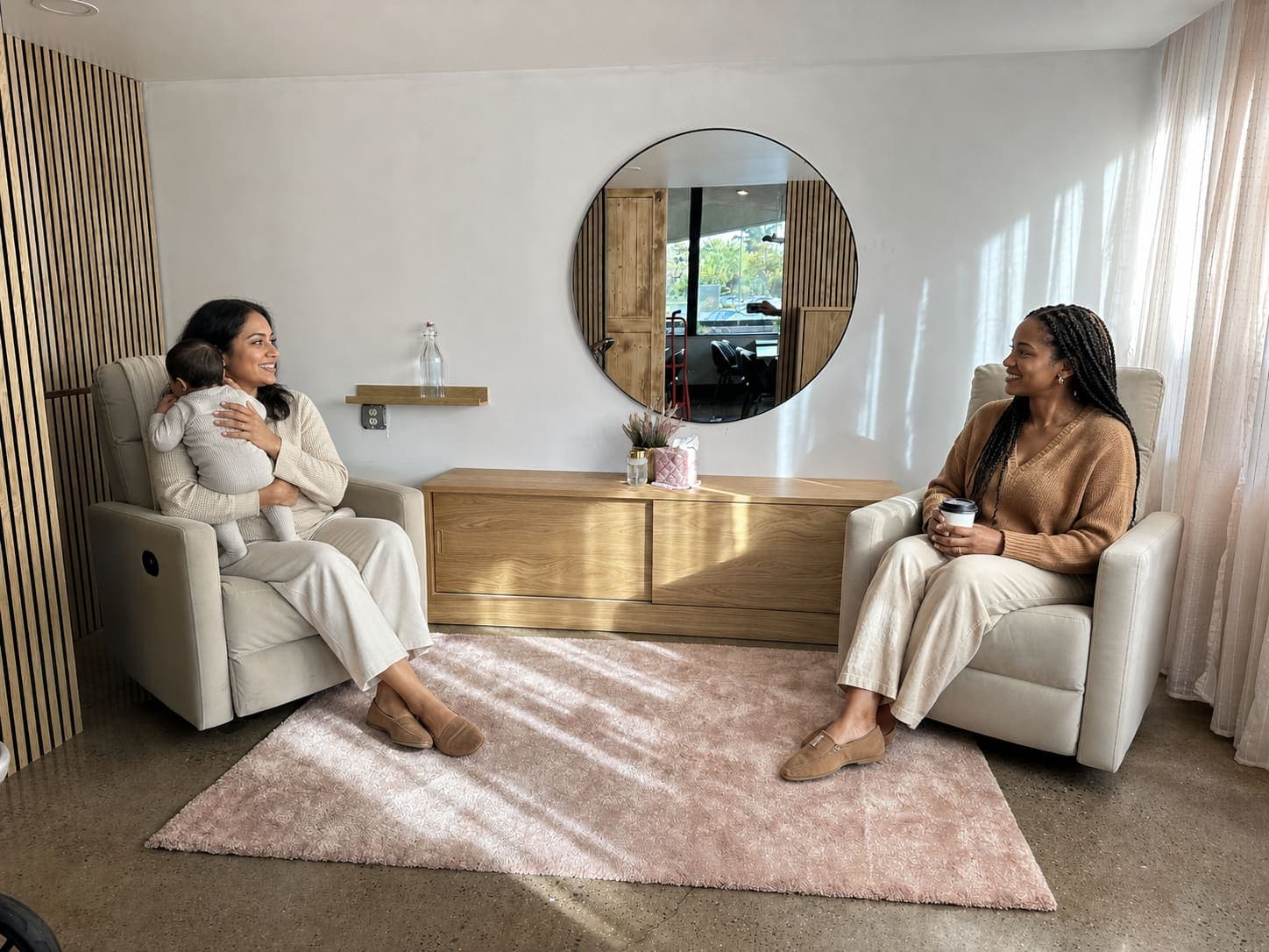 Two mamas relaxing in the recliners of the Mama Space, one holding her baby, the other holding a coffee, chatting across the rug.