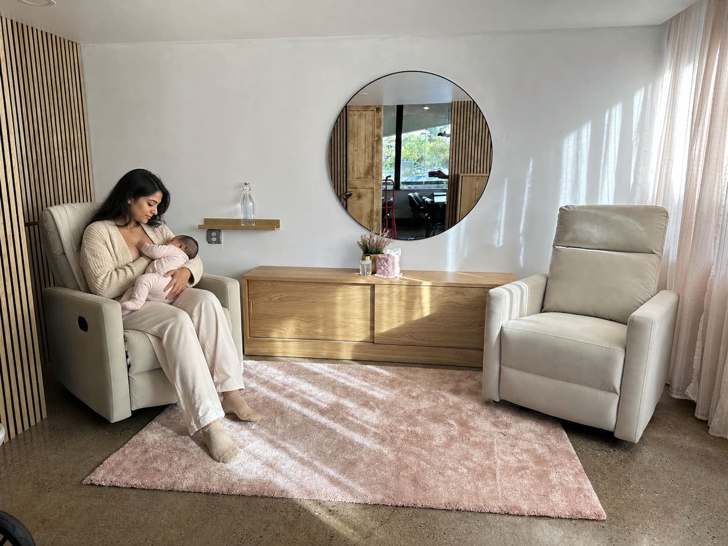 The Mama Space at iJava Willow Glen — a quiet private room with two plush recliners, a soft blush rug, a round mirror and a wood credenza, where a mama sits nursing her baby.