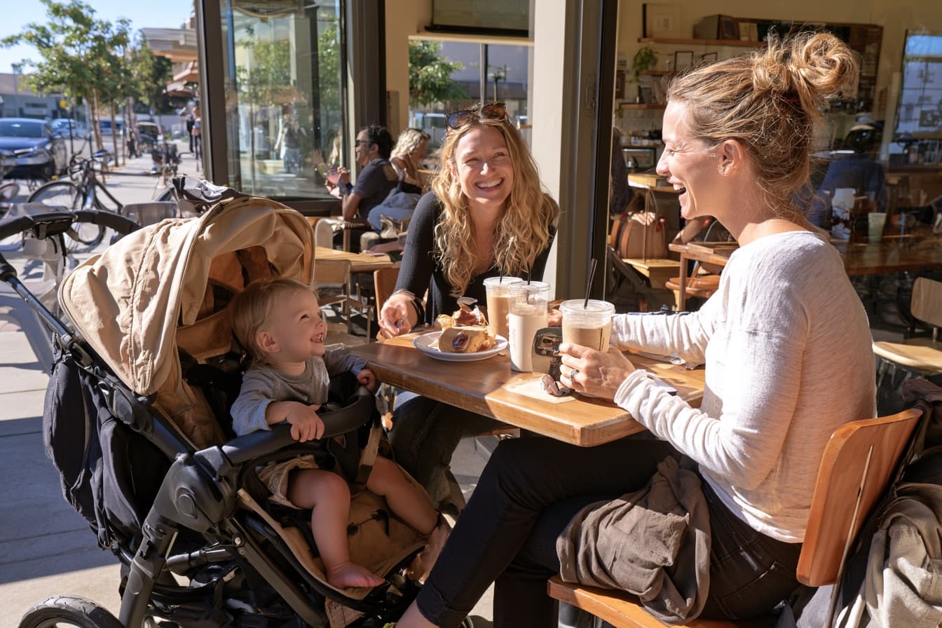 Two friends laughing over iced coffees on the sunny patio at iJava Willow Glen, a toddler smiling in a stroller beside them.