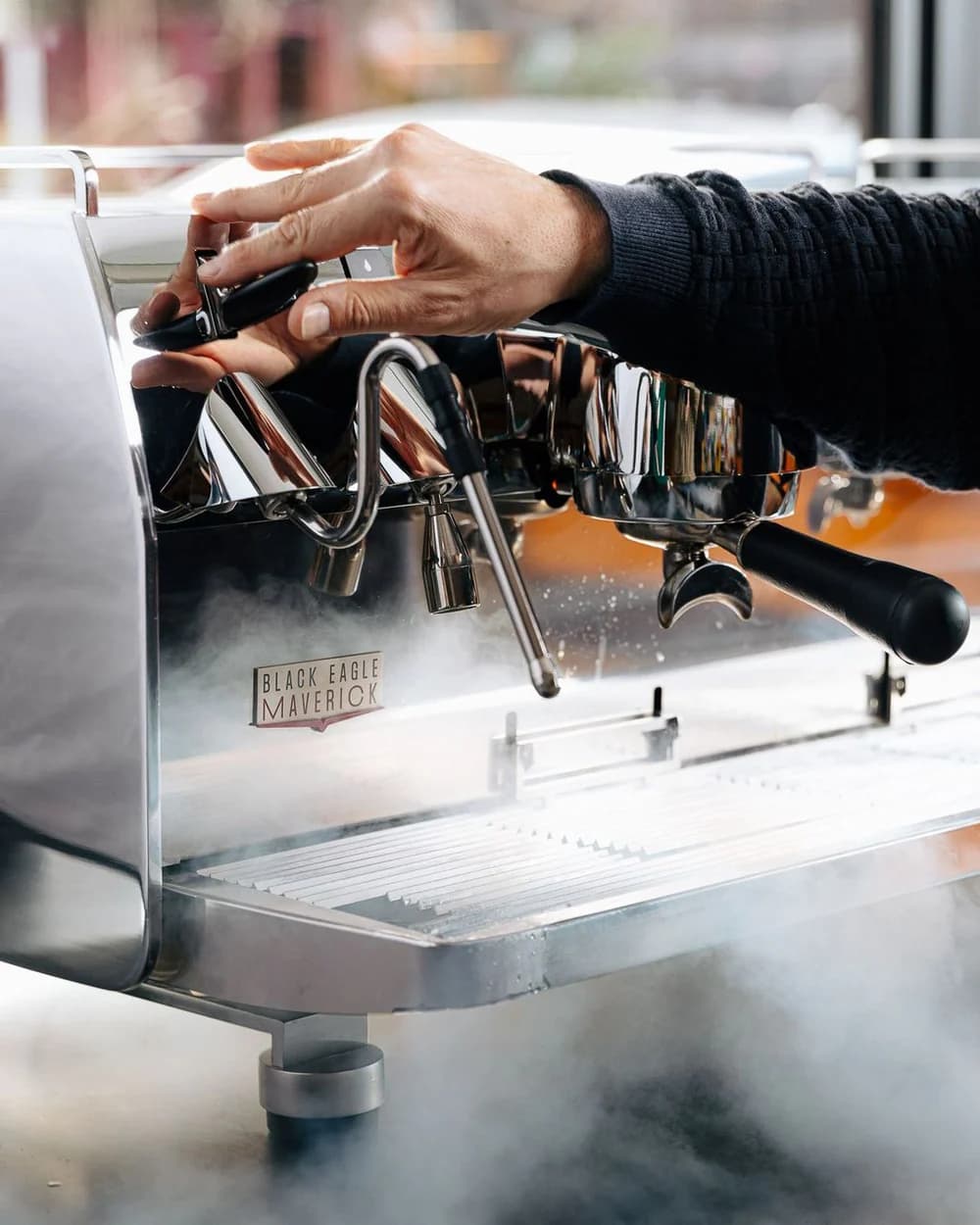 A hand adjusting the steam wand on a white Black Eagle Maverick, the badge visible beside a portafilter, steam rising.