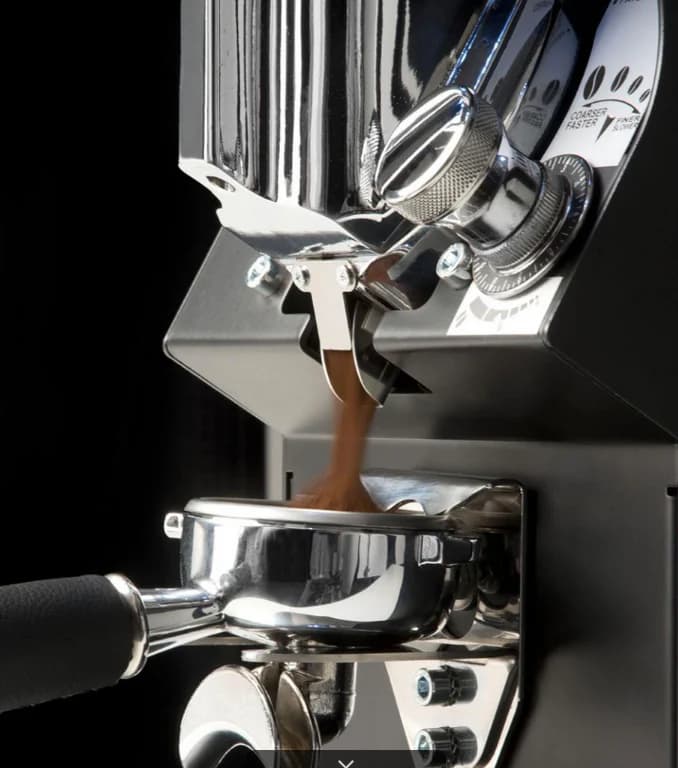 A Nuova Simonelli Mythos GX grinder dosing freshly ground coffee into a portafilter, grind-adjustment dial visible on its chrome body.